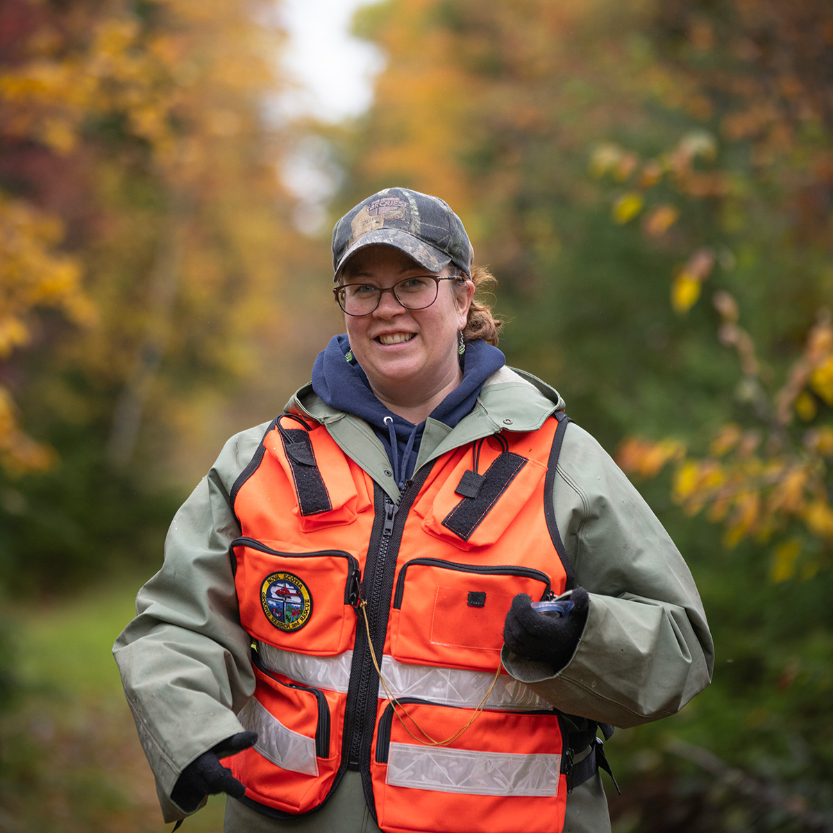 Smiling woman wearing a green rain coat, camouflage hat, and a reflective orange safety vest. She is holding a compass in her left hand.
