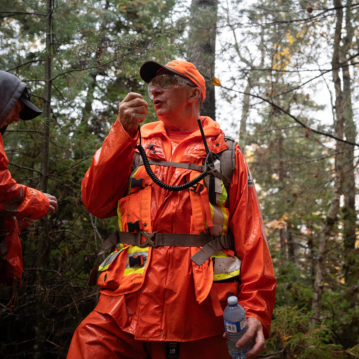 An older man speaking into a walkie talkie that is strapped to his side. He is wearing full orange and has a water bottle in his other hand.