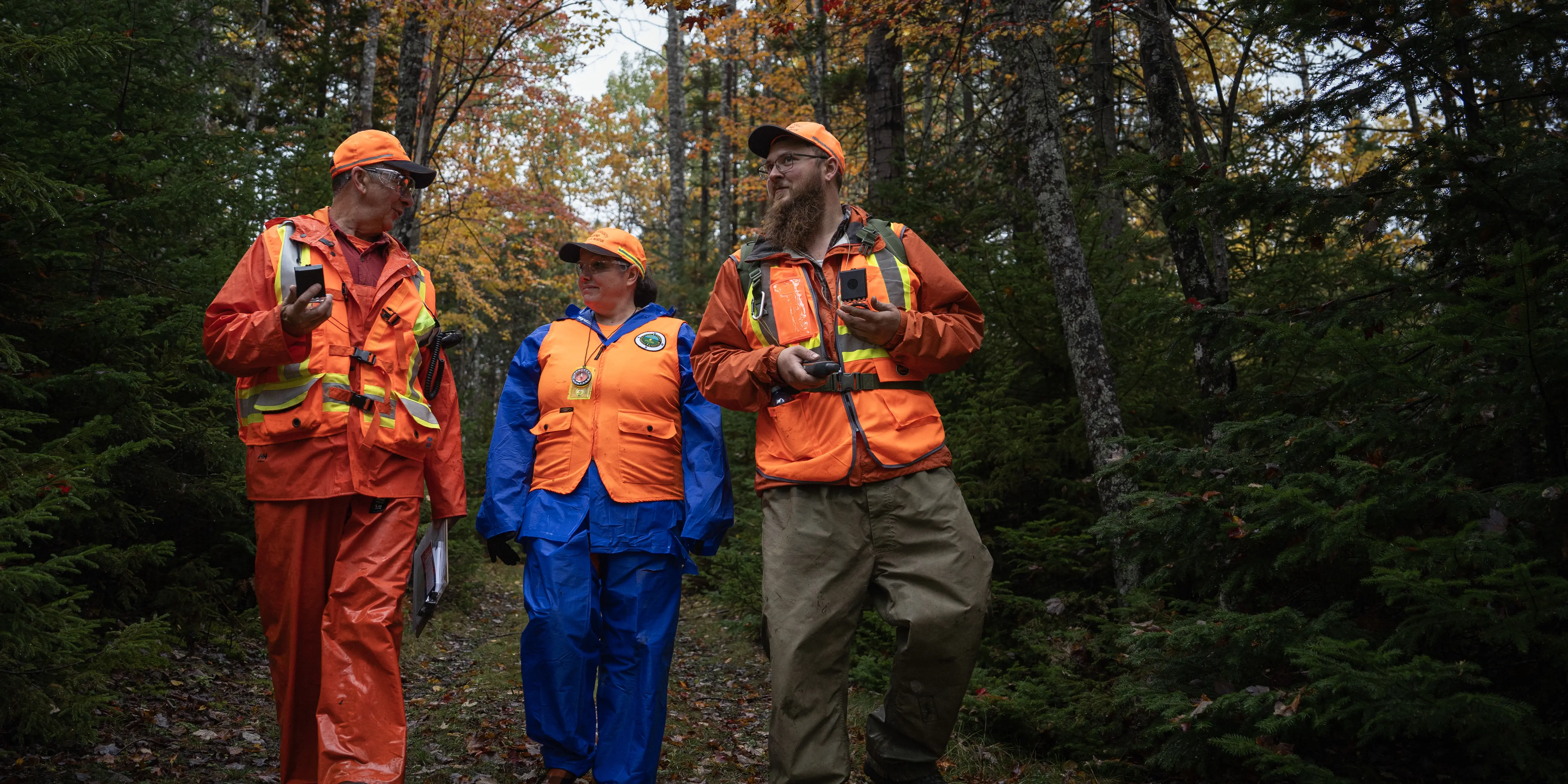 Three volunteers are smiling walking down a wooded trail. They each have a compass, one has a clipboard, and another has a GPS