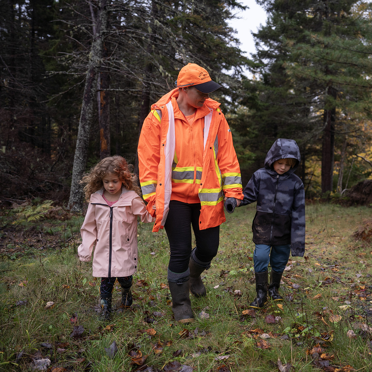 A woman wearing a bright reflective orange jacket and hat is holding two somber looking children's hands, walking out of the forest. The little girl on the left is wearing a pink jacket and the little boy is wearing blue.