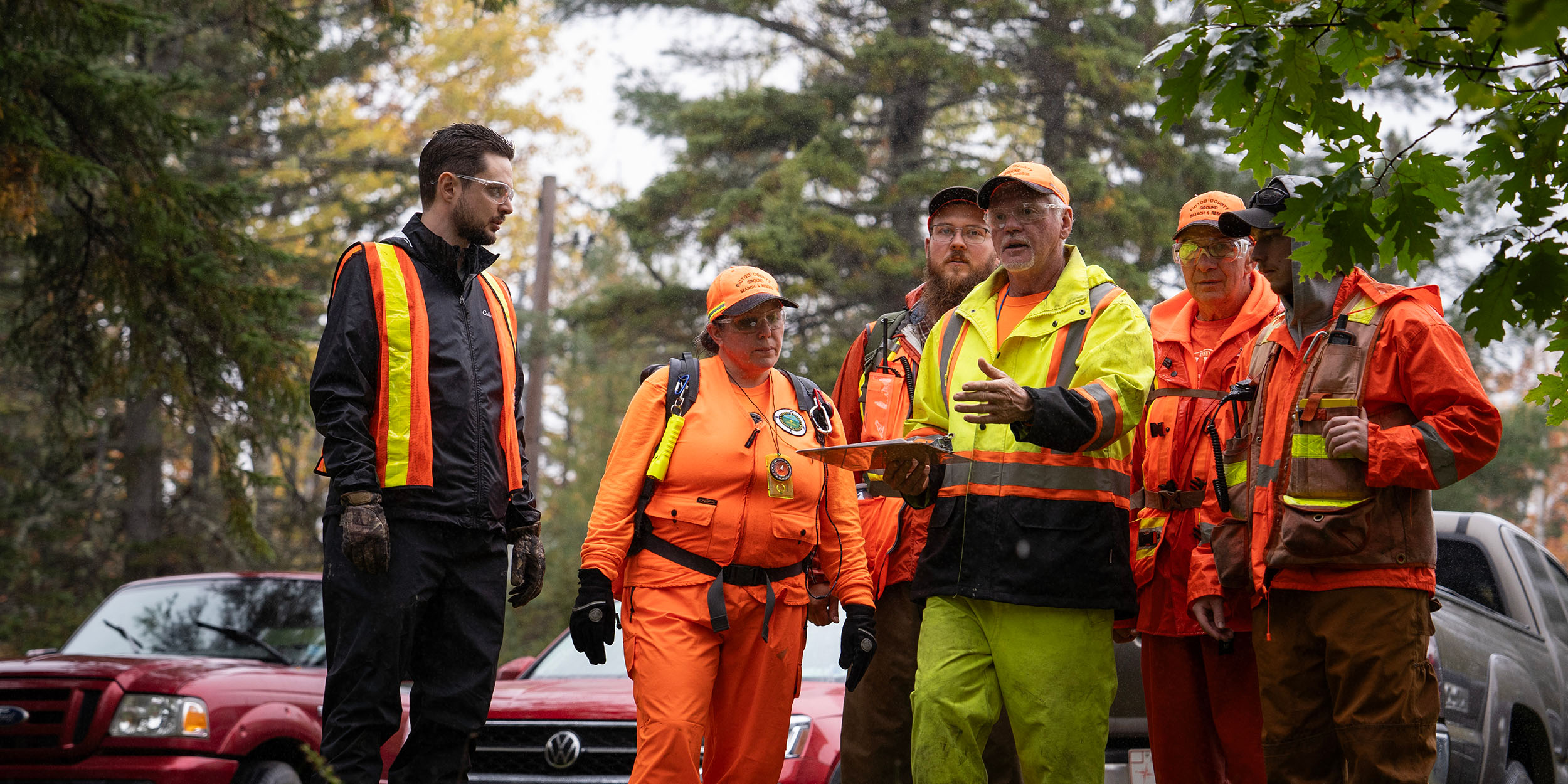 A group of five people stand around a sixth holding a clipboard, all listening intently.