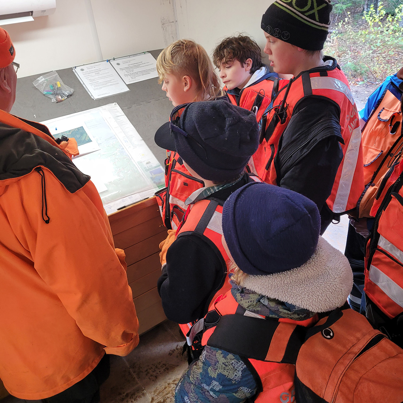 A group of children stand around a map as a volunteer searcher is pointing and explaining the information.